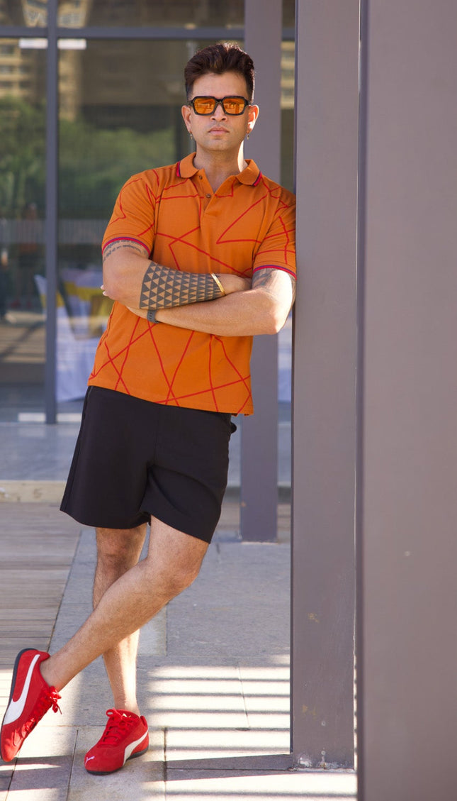 Man in orange shirt and black shorts standing in front of a glass wall with a cityscape reflection.