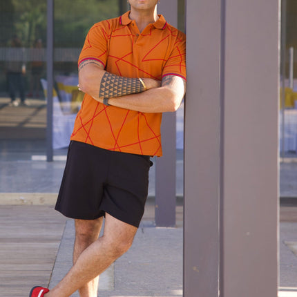 Man in orange shirt and black shorts standing in front of a glass wall with a cityscape reflection.
