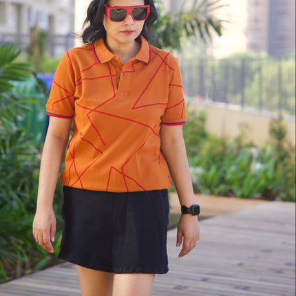 Woman wearing an orange polo shirt and black skirt standing on a wooden deck with buildings and greenery in the background.