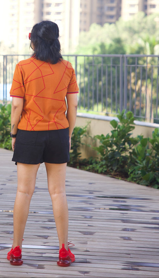 Person wearing an orange shirt and black shorts standing on a wooden deck with buildings and greenery in the background