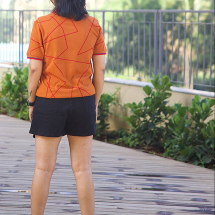 Person wearing an orange shirt and black shorts standing on a wooden deck with buildings and greenery in the background