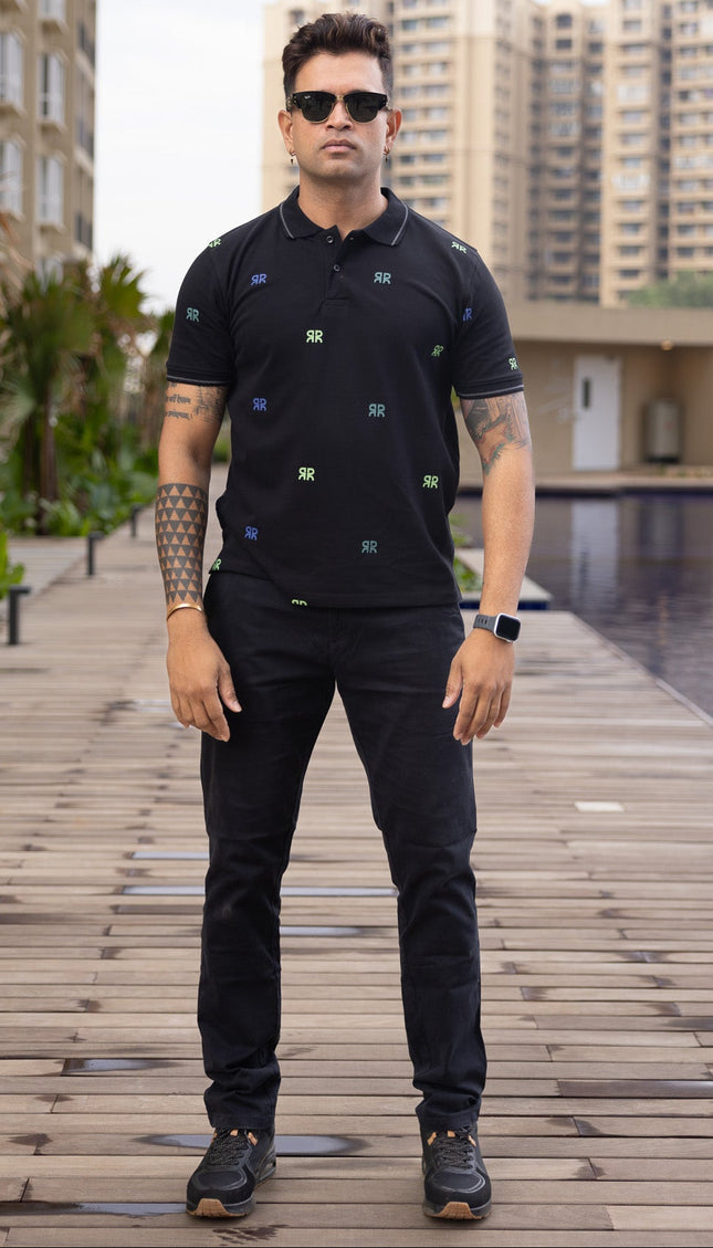 Man in black outfit with sunglasses standing on a wooden pathway by a pool with buildings in the background