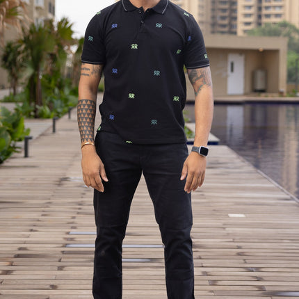 Man in black outfit with sunglasses standing on a wooden pathway by a pool with buildings in the background