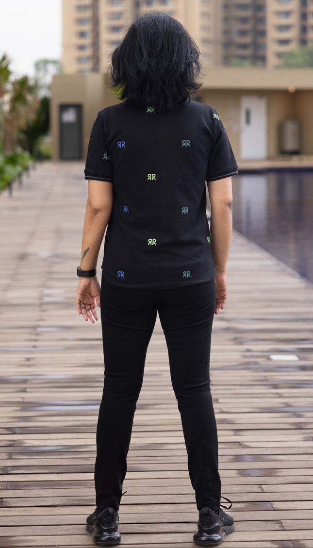 Person wearing a black t-shirt with small logos standing on a wooden pathway with buildings and a pool in the background.