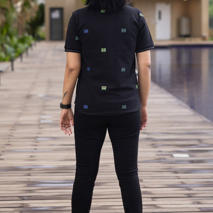 Person wearing a black t-shirt with small logos standing on a wooden pathway with buildings and a pool in the background.
