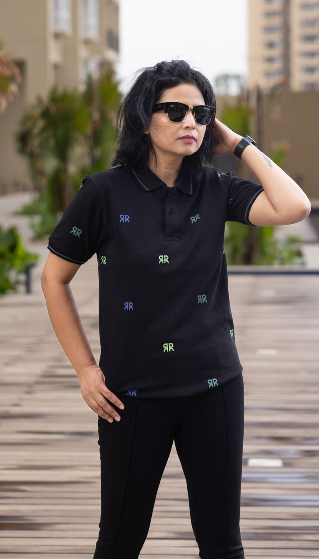Person wearing a black polo shirt with small patterns, standing on a wooden deck with buildings in the background.