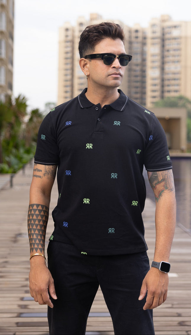 Man wearing a black polo shirt with small colorful patterns, standing by a pool with buildings in the background.