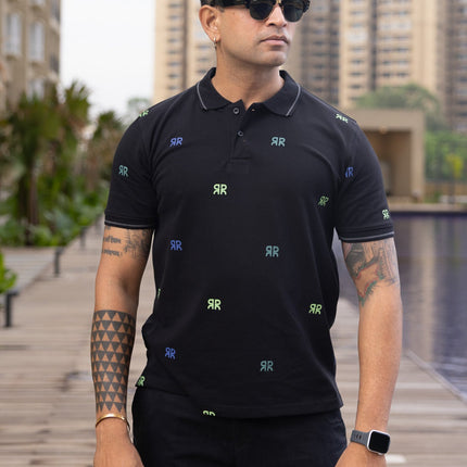 Man wearing a black polo shirt with small colorful patterns, standing by a pool with buildings in the background.
