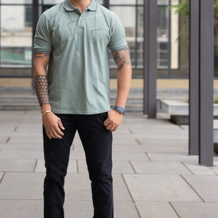 Man wearing a green polo shirt and dark pants standing under a wooden canopy.