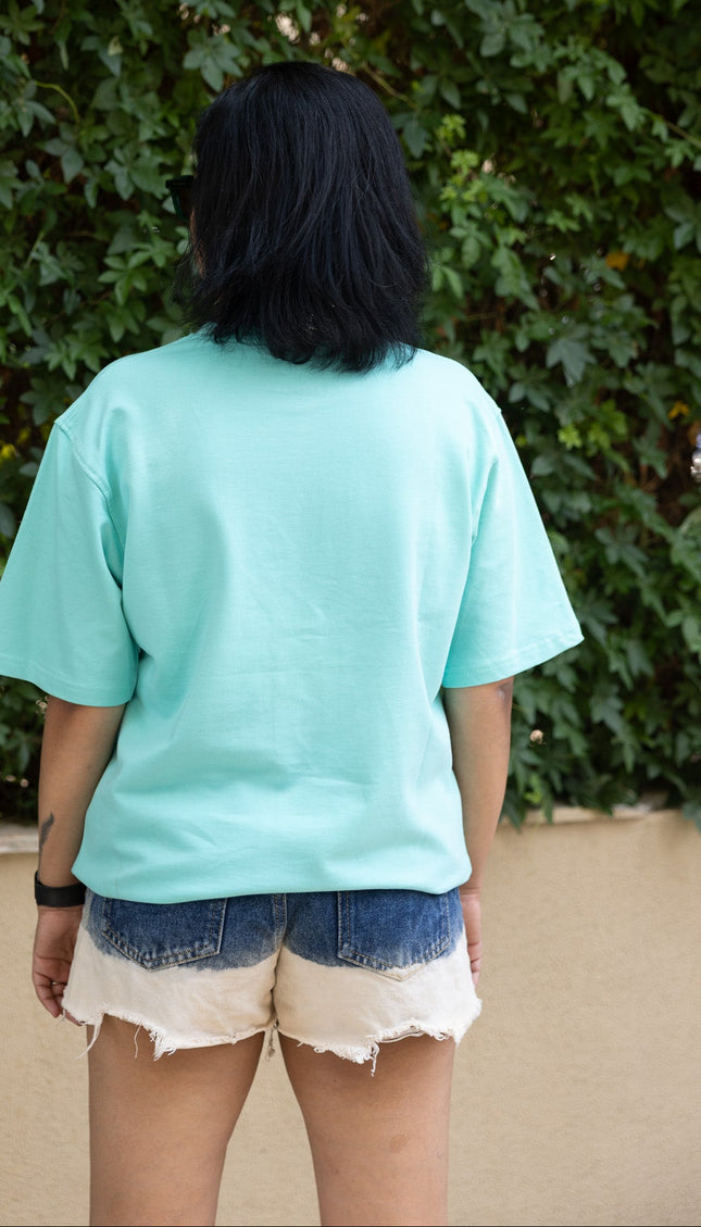 Person wearing a light blue t-shirt and denim shorts standing in front of green foliage.