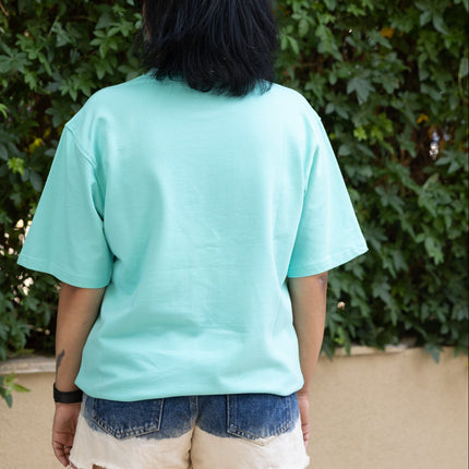 Person wearing a light blue t-shirt and denim shorts standing in front of green foliage.
