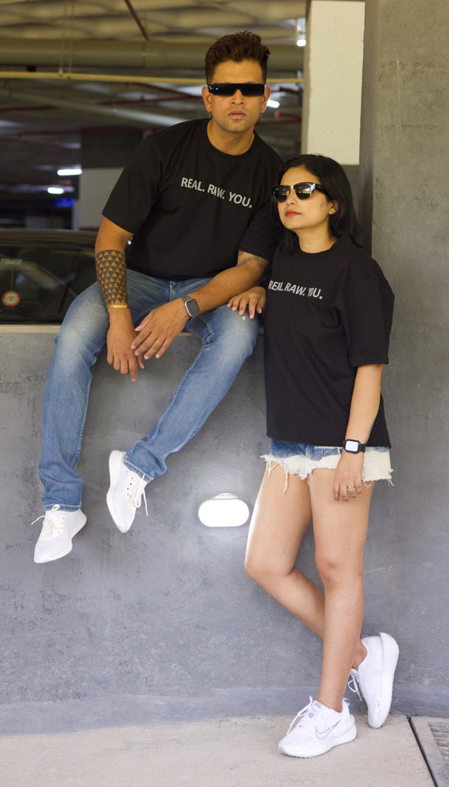 Two people sitting on a concrete floor in an indoor setting, wearing black t-shirts with text.