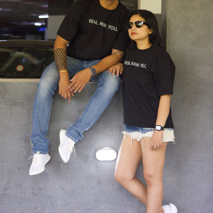 Two people sitting on a concrete floor in an indoor setting, wearing black t-shirts with text.