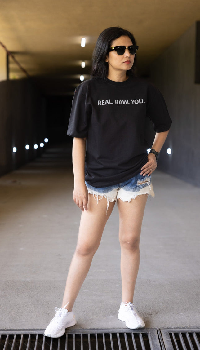 Person wearing a black t-shirt with text, denim shorts, and white sneakers in an urban setting.