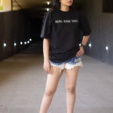 Person wearing a black t-shirt with text, denim shorts, and white sneakers in an urban setting.
