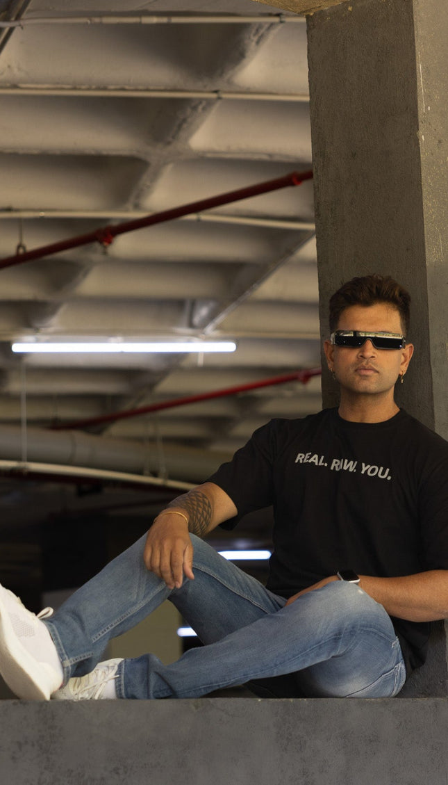 Man sitting on a concrete ledge wearing sunglasses and a black t-shirt with text.