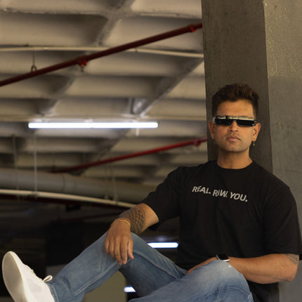 Man sitting on a concrete ledge wearing sunglasses and a black t-shirt with text.