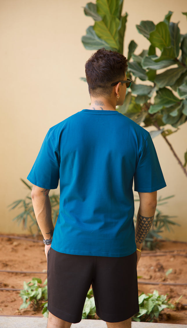 Person wearing a blue shirt and black shorts standing in front of a plant with a beige wall background