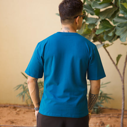 Person wearing a blue shirt and black shorts standing in front of a plant with a beige wall background