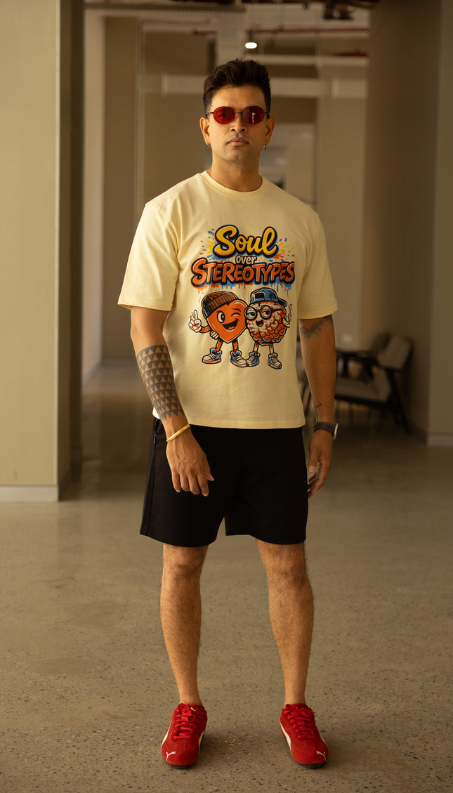 Man wearing a beige t-shirt with a graphic design, black shorts, and red shoes in an indoor setting.