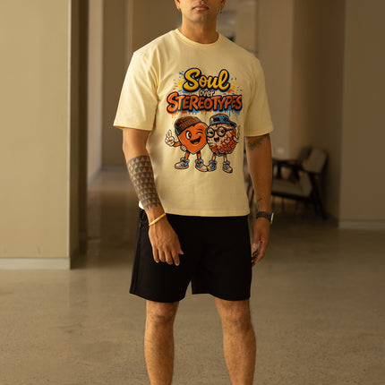 Man wearing a beige t-shirt with a graphic design, black shorts, and red shoes in an indoor setting.