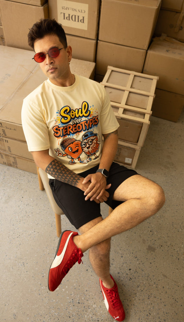 Man sitting on a chair wearing a graphic t-shirt and red shoes in a room with cardboard boxes.