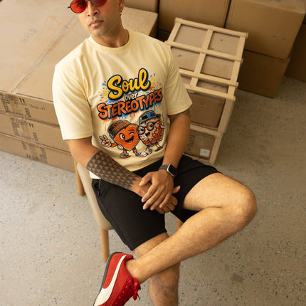 Man sitting on a chair wearing a graphic t-shirt and red shoes in a room with cardboard boxes.